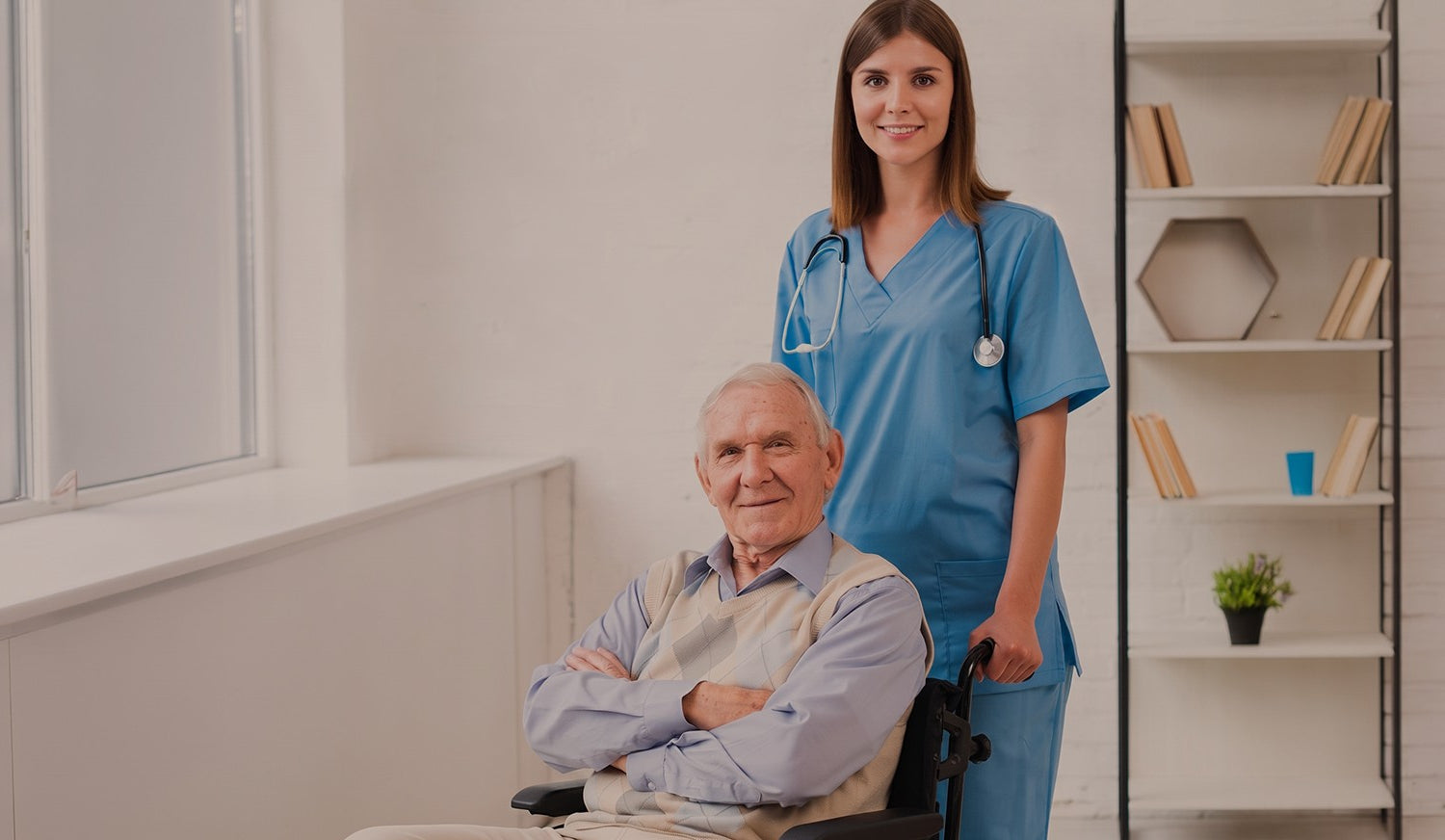 Enfermera o cuidadora profesional en uniforme azul sonriendo junto a un adulto mayor en silla de ruedas en un entorno luminoso.