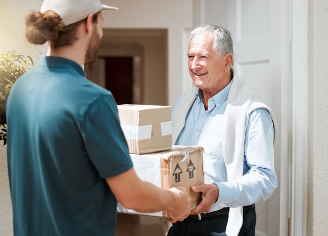 Entrega a domicilio de productos para adultos mayores en El Salvador: Un repartidor de Juby Care entrega paquetes a un hombre mayor sonriente en la puerta de su hogar, destacando el servicio de delivery seguro y confiable para la comodidad de los grandes de la casa.