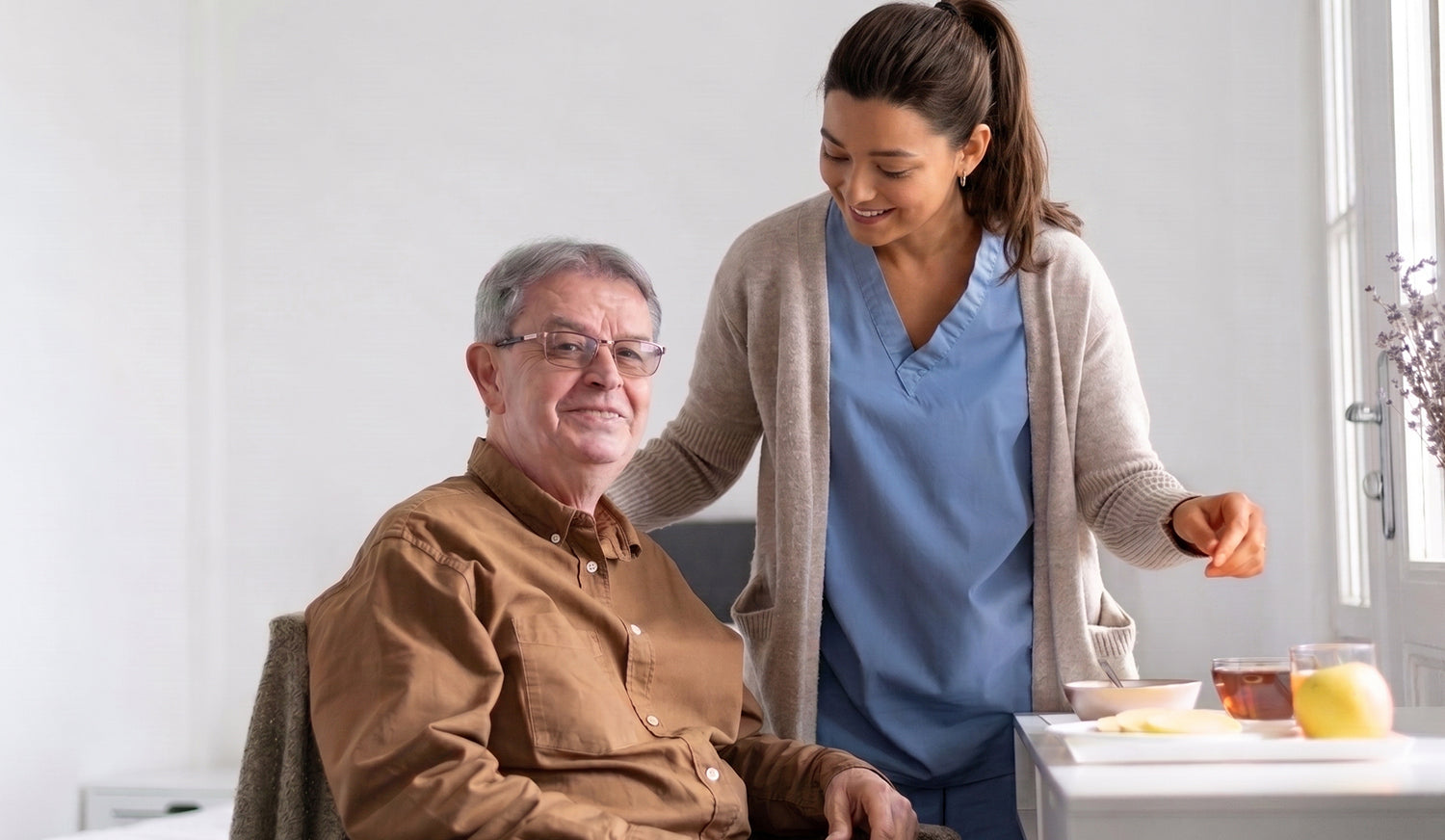Una enfermera o cuidadora sonriente con uniforme médico sirve un desayuno saludable con fruta a un adulto mayor sentado en su comedor.