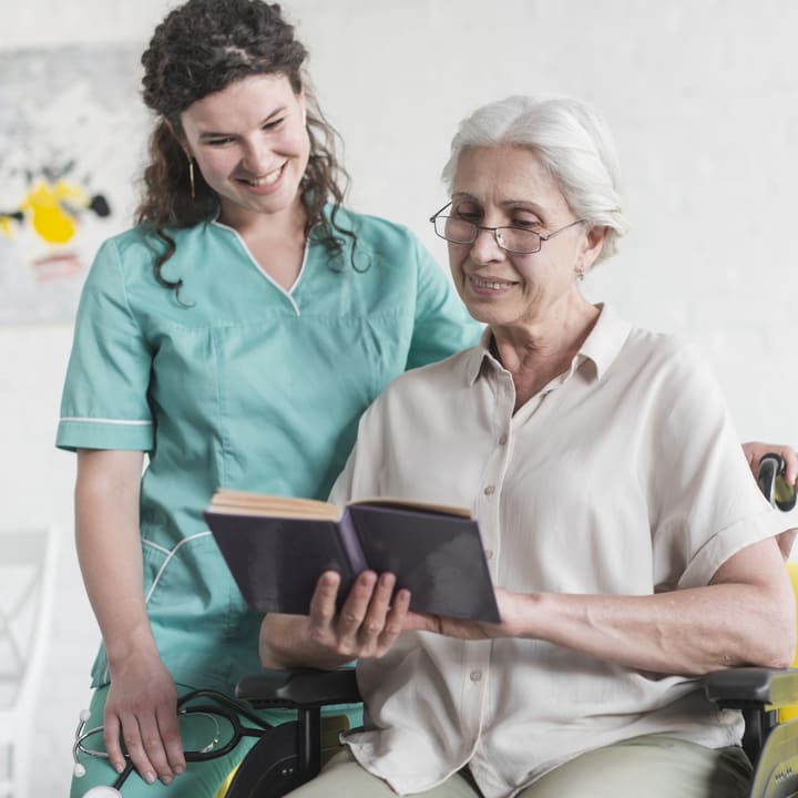 Acompañamiento integral y cuidado especializado para adultos mayores en El Salvador: una integrante del Juby Care Team comparte un momento de lectura con una mujer de la tercera edad en silla de ruedas, brindando apoyo emocional con respeto y cercanía humana para garantizar su bienestar y dignidad.